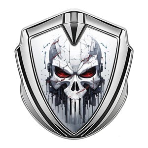 May include: A silver shield-shaped emblem featuring a detailed skull design with red eyes and a cracked, white head. The skull has a mechanical appearance, with dark drips and accents.