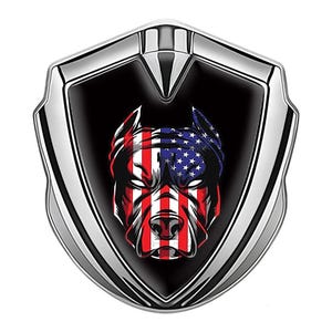 May include: A shield-shaped emblem with a silver border and a black center. The center features a stylized dog's head with the American flag design, using red and white stripes and blue stars.