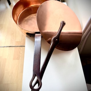 May include: A copper pan with a matching lid. The pan has a rounded shape with a wide opening and a dark handle. The lid is oval and has a dark handle. The pan and lid are sitting on a white surface.