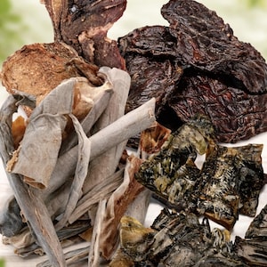 以下が含まれることがあります： さまざまな乾燥ペット用おやつのクローズアップ。濃い茶色のジャーキー、ベージュとグレーのストリップ、緑がかった部分が含まれています。おやつは山のように配置され、さまざまなテクスチャと色を示しています。