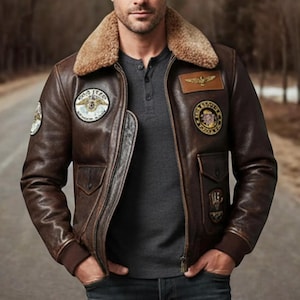 Puede incluir: Chaqueta de cuero marrón con cuello de borrego y parches bordados. La chaqueta tiene cremallera, dos bolsillos delanteros y puños de canalé. Se lleva sobre una camiseta gris de manga larga.