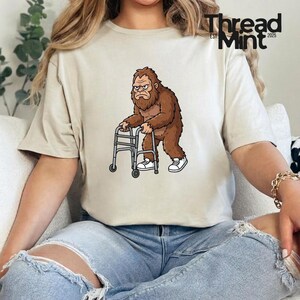 May include: Beige t-shirt featuring a cartoon Bigfoot character with a grumpy expression, using a walker. The Bigfoot is brown with white sneakers. The shirt has the text "Thread Mint EST. 2025" in black.
