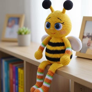 May include: A handmade, crocheted bee doll with a yellow body, black stripes, and large blue eyes. The bee has white wings, black antennae, and striped arms and legs. It is sitting on a wooden shelf.