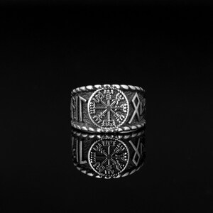 Puede incluir: Un anillo plateado con un diseño detallado. El anillo presenta un símbolo central de rosa de los vientos, rodeado de runas y un borde trenzado. El anillo se muestra sobre una superficie negra reflectante.