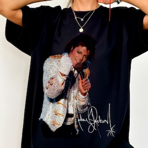 May include: Black t-shirt featuring a vibrant image of Michael Jackson in a performance pose, holding a microphone. The image includes his signature in white. The shirt is a classic crew neck style.