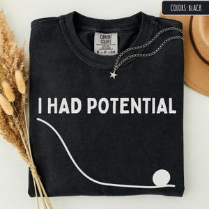 May include: Black t-shirt with the text "I HAD POTENTIAL" in white, along with a white graphic of a curved line and a circle. A silver necklace with a star pendant rests on the shirt. The shirt has a Comfort Colors tag.