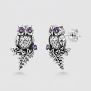 Op de afbeelding: Een paar zilveren uilenoorknopjes. Elke uil zit op een tak met varenbladeren en kleine paarse edelstenen. De ogen van de uil zijn ook paars. Kleine witte edelstenen bevinden zich ook op de tak.