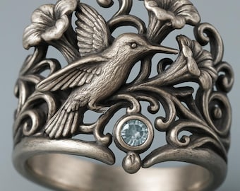 Könnte beinhalten: Silberfarbener Ring mit einem fliegenden Kolibri, umgeben von stilisierten Blumen und Blättern. Der Ring hat ein aufwendiges, detailliertes Design mit einem kleinen, runden, hellblauen Edelstein. Das Design des Rings ist von der Natur inspiriert.