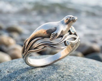 May include: A silver ring featuring a sculpted seal resting on a band designed to resemble flowing water. The band incorporates two small, round, light blue gemstones set into the metal waves, creating a detailed and cohesive aquatic motif.
