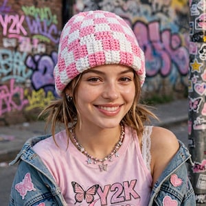 May include: A crochet beanie featuring a pink and white checkered pattern. The hat is constructed with a thick yarn texture and a rounded crown shape that fits snugly on the head, showcasing a consistent grid design throughout the entire piece.