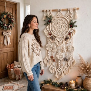 Op de afbeelding: Een grote crèmekleurige macramé wandhanger met een centraal cirkelvormig web, bungelende spiraalvormen, een ster, een kleine boom, dennenappels, kaneelstokjes en takjes groen met rode bessen, opgehangen aan een houten roede.