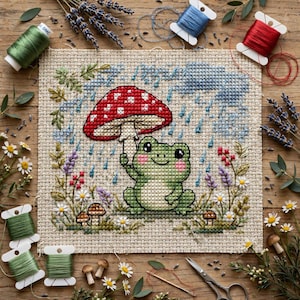 May include: A cross-stitch embroidery piece on beige fabric featuring a green frog holding a red and white spotted mushroom as an umbrella. The scene includes blue rain stitches, small brown mushrooms, and colorful floral accents. Spools of green, blue, and red thread, lavender sprigs, and metal scissors are arranged around the fabric.