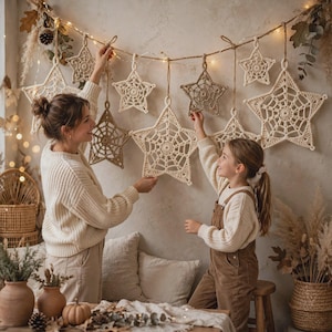 May include: A decorative garland featuring multiple crocheted stars in varying sizes and shades of cream and tan. The stars are suspended from a string adorned with small warm white fairy lights, creating a festive display of intricate yarn work.
