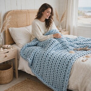 May include: A chunky knit throw blanket in a light blue color featuring a thick, oversized yarn texture. The blanket is draped over a bed, showcasing its heavy, soft, and interconnected loop construction that provides a dense and cozy appearance.