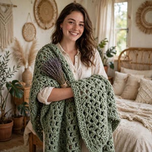 May include: A sage green chunky knit throw blanket with a large open-weave pattern, draped over the arms and held alongside a dried bundle of purple lavender stems. The blanket features thick yarn loops and a textured, soft appearance throughout.