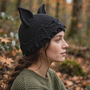 May include: A black crocheted beanie featuring two prominent pointed ears on top and a decorative bat wing pattern integrated into the lower edge of the hat. The design includes a small, detailed bat motif centred above the forehead area.