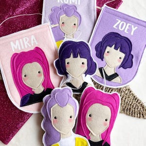 May include: Felt banner decorations with illustrated faces and names. The banners are in shades of pink and purple, with white text that reads "MIRA" and "ZOEY". The faces have blush-coloured cheeks and various hairstyles.