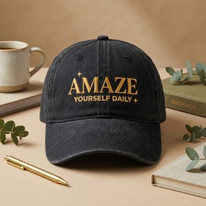 May include: A black baseball cap with the word "AMAZE" embroidered in gold, along with "YOURSELF DAILY +." The cap is made of a soft, worn material. A gold pen, a mug, and a book are in the background.