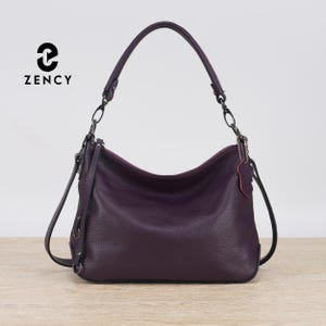 Puede incluir: Bolso de hombro de cuero morado oscuro con diseño curvo. Cuenta con un asa superior y una correa de hombro desmontable. El bolso tiene un cierre de cremallera visible y una pequeña etiqueta de cuero. La marca "ZENCY" está impresa en la parte superior izquierda.