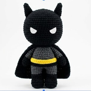 Puede incluir: Muñeco de Batman hecho a mano con ganchillo. El muñeco es principalmente negro con un torso y piernas grises. Presenta un cinturón amarillo, ojos blancos y orejas puntiagudas. El muñeco tiene una capa negra y está de pie.