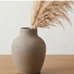 Puede incluir: Un jarrón de cerámica beige texturizado, con un cuerpo redondeado y un cuello estrecho. El jarrón contiene un manojo de hierba de la pampa seca y plumosa. El jarrón está sobre una superficie de madera, creando una estética neutra y terrosa.