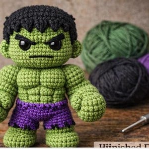 Könnte beinhalten: Handgefertigte Hulk-Puppe aus Häkelarbeit mit grünem Körper, schwarzem Haar und lila Shorts. Die Puppe hat einen wütenden Gesichtsausdruck und steht. Grün, schwarze und lila Wollknäuel befinden sich im Hintergrund.