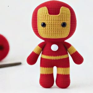 May include: A handmade crochet Iron Man doll in red and yellow. The doll has a round head with a yellow mask, black eyes, and a white circle on the chest. A red ball and a crochet hook are also in the image.