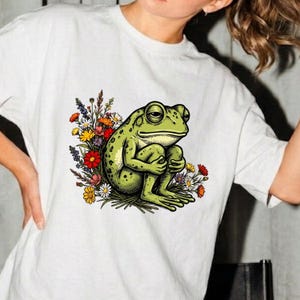May include: A white short-sleeved t-shirt featuring a graphic illustration of a green frog sitting in a cluster of colorful wildflowers. The frog is depicted with a textured skin pattern, and the surrounding flowers include red, yellow, and white blossoms.