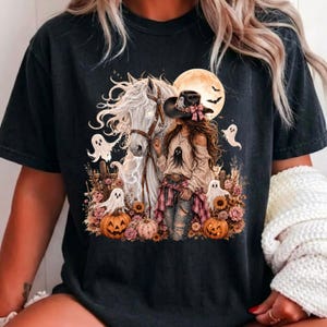 May include: A black t-shirt featuring a graphic print of a person in a cowboy hat standing next to a white horse. The design includes a full moon, flying bats, small ghosts, pumpkins, and floral elements in shades of orange, pink, and brown.