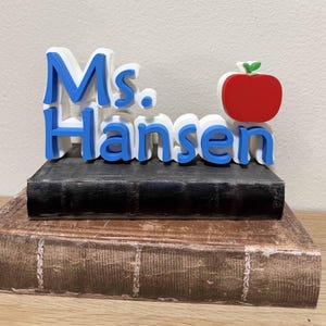 May include: A teacher-themed desk decoration featuring the name "Ms. Hansen" in blue and white 3D lettering, topped with a red apple. The lettering sits atop a black book, which rests on a vintage-looking book.