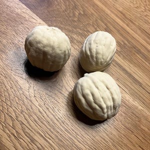 May include: Three off-white, textured, spherical objects with a ribbed surface are arranged on a wooden surface. The objects have a natural, organic appearance, resembling small, stylized nuts or seed pods. The wood grain provides a warm, contrasting backdrop.