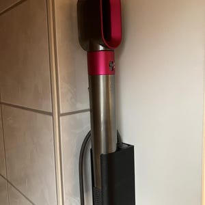 Könnte beinhalten: Ein Dyson-Haartrockner in einer schwarzen Wandhalterung. Der Haartrockner ist silberfarben mit einem rosa und dunkelgrauen Kopf. Die Halterung ist an einer weißen Wand mit grauen Fliesen befestigt.