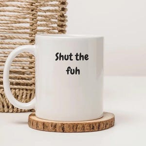 May include: White ceramic mug with the text "Shut the fuh" in black. The mug is on a round wooden coaster. A woven basket is in the background.