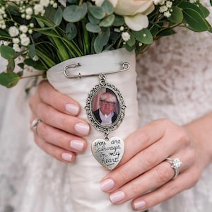 Op de afbeelding: Een bruidsboeket met roze en witte rozen, groen en een herdenkingsbedel. De bedel heeft een foto in een ovale lijst en een hartvormige hanger met de tekst "You are always in my heart!"