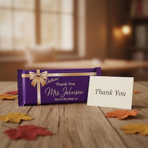May include: A purple Cadbury chocolate bar wrapper featuring a gold ribbon graphic, the text Thank You Mrs. Johnson You're the Best! xx, and a small white folded card with the text Thank You printed in black on the front of the card.