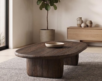 May include: A low, oval-shaped coffee table crafted from dark, textured wood with a thick, rounded top and two sturdy, block-like pedestal bases. A small, shallow, light-colored bowl sits centered on the dark wooden surface.