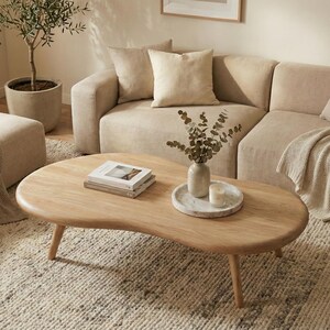 May include: A modern living room setup featuring a beige sectional sofa, a wooden coffee table, and a textured rug. Decorative pillows and a throw blanket add to the cozy atmosphere. A potted olive tree and a vase with greenery complete the scene.