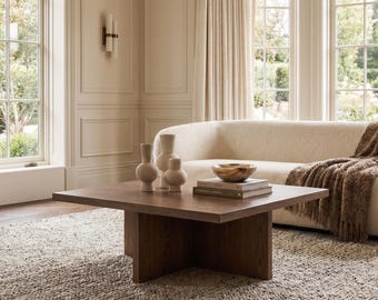 May include: A square dark brown wooden coffee table with a pedestal base, featuring three light-colored ceramic vases and a small bowl resting on a stack of books. A cream-colored curved sofa with a brown textured throw blanket sits nearby.