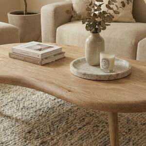 May include: A kidney-shaped light oak coffee table with three rounded legs. On the surface sits a stack of two books titled Kinfolk, a white marble circular tray, a light-colored ceramic vase with dried foliage, and a small glass candle.