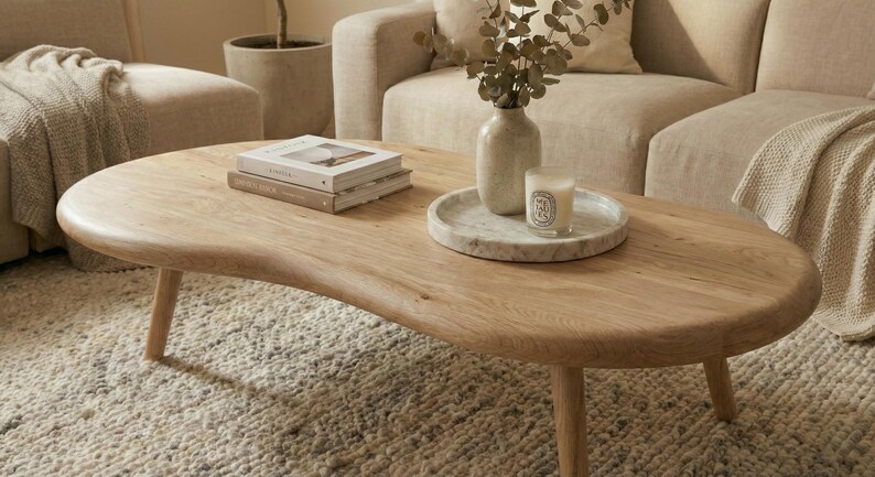 May include: A kidney-shaped light oak coffee table with three rounded legs. On the surface sits a stack of two books titled Kinfolk, a white marble circular tray, a light-colored ceramic vase with dried foliage, and a small glass candle.