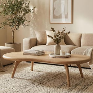 May include: A light wood coffee table with an oval top and four tapered legs. The table is positioned in front of a beige sofa with matching throw blankets and pillows, featuring a small vase and books on its surface in a bright living room.