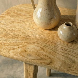 K&ouml;nnte beinhalten: Ein heller Beistelltisch aus Holz mit abgerundeten Ecken und sichtbarer Maserung. Auf der Tischoberfl&auml;che stehen drei Keramikgef&auml;&szlig;e: ein gro&szlig;er gesprenkelter Krug, eine kleine runde Vase und ein hoher, weit ge&ouml;ffneter gesprenkelter Becher.