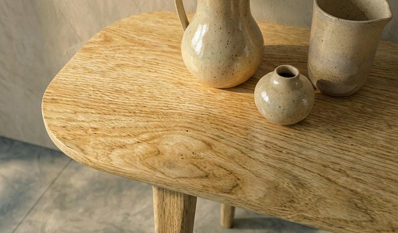 K&ouml;nnte beinhalten: Ein heller Beistelltisch aus Holz mit abgerundeten Ecken und sichtbarer Maserung. Auf der Tischoberfl&auml;che stehen drei Keramikgef&auml;&szlig;e: ein gro&szlig;er gesprenkelter Krug, eine kleine runde Vase und ein hoher, weit ge&ouml;ffneter gesprenkelter Becher.
