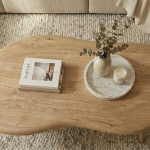 May include: A light brown wooden coffee table with an irregular, organic shape. On the surface sits a stack of books with the word Kinfolk visible on the cover, a round white marble tray, a beige ceramic vase with eucalyptus, and a small candle.