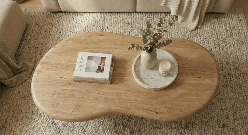 May include: A light brown wooden coffee table with an irregular, organic shape. On the surface sits a stack of books with the word Kinfolk visible on the cover, a round white marble tray, a beige ceramic vase with eucalyptus, and a small candle.