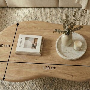 May include: A light brown wooden coffee table with an irregular organic shape. The table measures 120 inches by 60 inches. On the surface sits a book titled Kinfolk, a white marble tray, a ceramic vase with eucalyptus, and a small candle.