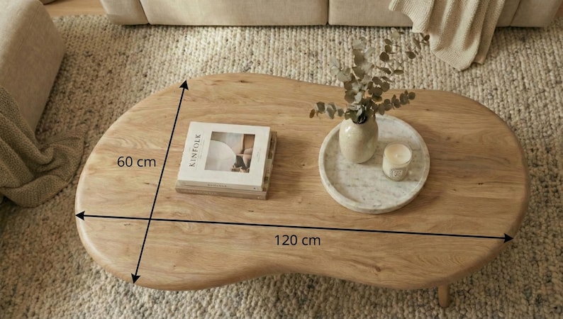 May include: A light brown wooden coffee table with an irregular organic shape. The table measures 120 inches by 60 inches. On the surface sits a book titled Kinfolk, a white marble tray, a ceramic vase with eucalyptus, and a small candle.