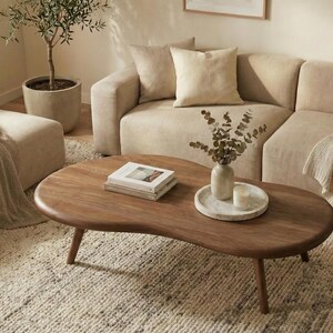 May include: A kidney-shaped wooden coffee table with a smooth, dark brown finish and three tapered legs. The table surface holds a stack of two books, a small ceramic vase containing dried eucalyptus stems, and a white circular tray with a candle.