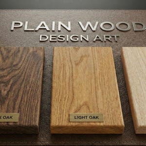 May include: A display board titled PLAIN WOOD DESIGN ART featuring three rectangular wood samples. The samples are labeled with small metal plates reading DARK OAK, LIGHT OAK, and NATURAL, showcasing distinct wood grain patterns and color tones.