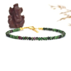 Puede incluir: Una delicada pulsera con pequeñas cuentas facetadas verdes y rubí ensartadas. La pulsera tiene un cierre dorado y se muestra sobre un fondo blanco. Una pequeña estatua marrón está en el fondo.